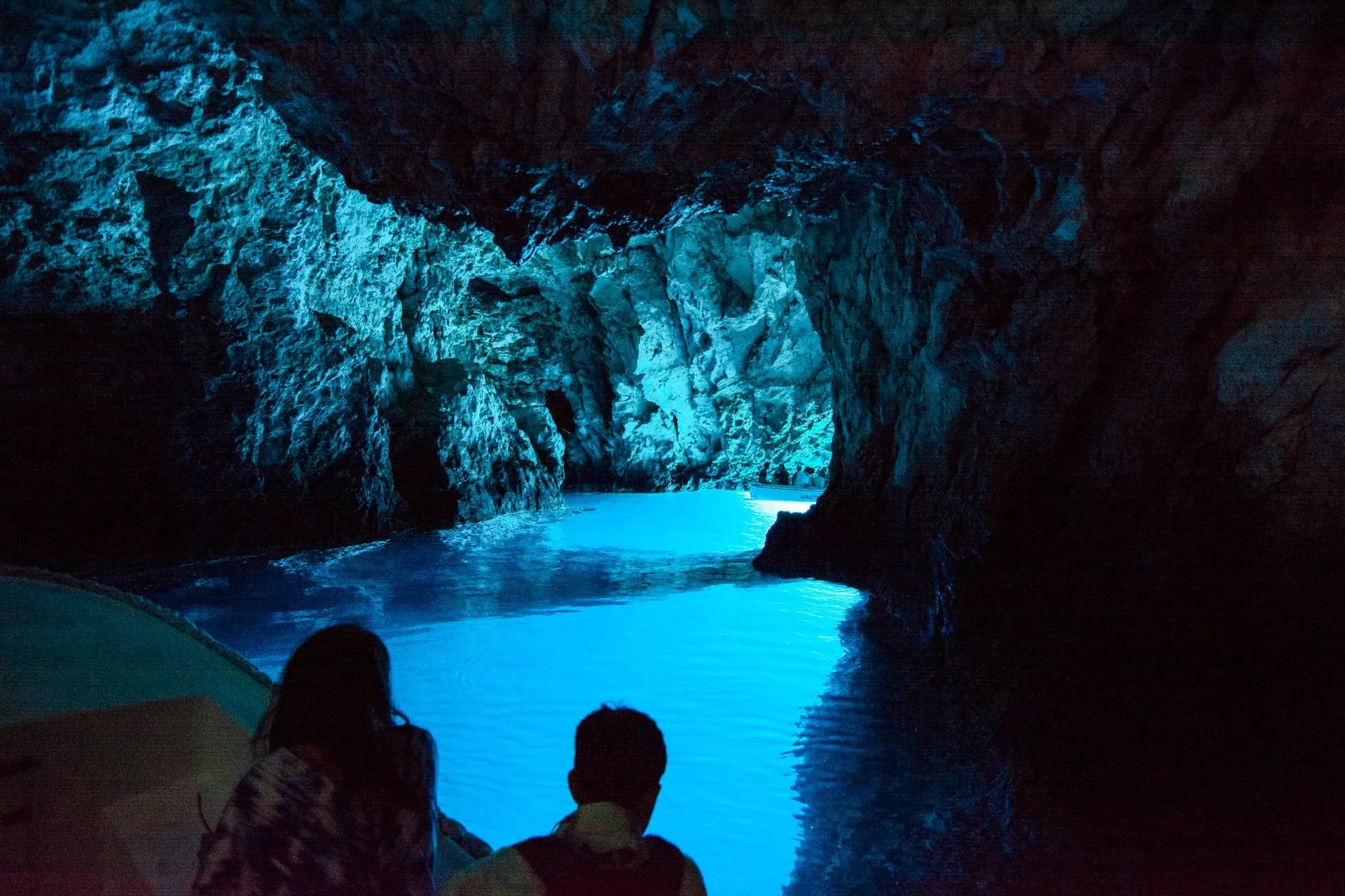 Inside the Blue Cave on Biševo from the boat, guests in silhouette against water lit that electric blue from the opening below the surface. Blue Cave & 5 Islands: Private
